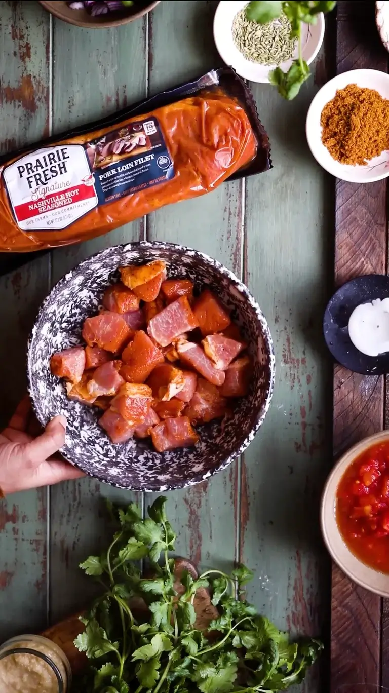 Cubed pieces of Prairie Fresh® Nashville Hot pork loin filet in a bowl surrounded by spices and fresh herbs.